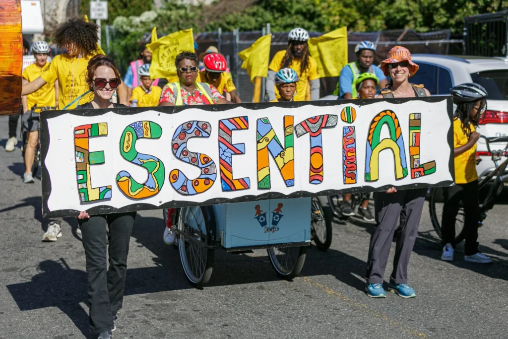 People and bicyclists in a parade holding up a banner that says "Essential"