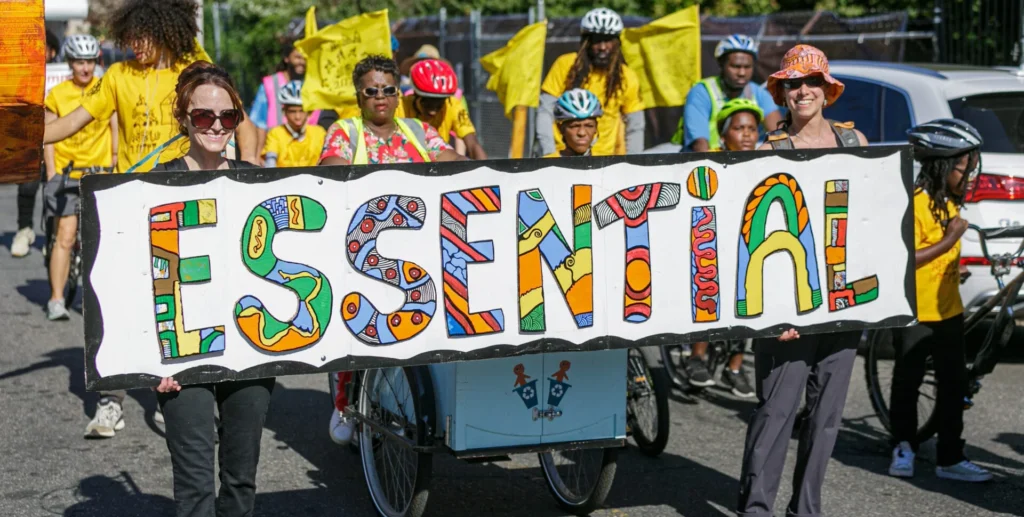 People and bicyclists in a parade holding up a banner that says "Essential"