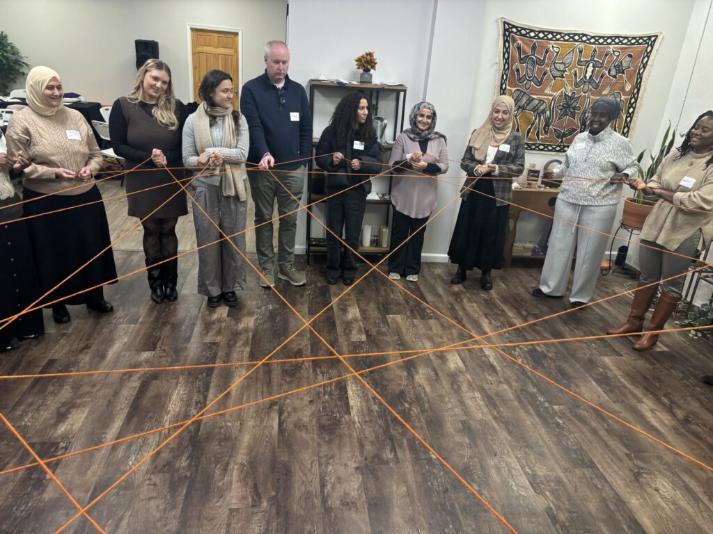 A group of people in a circle hold string that makes a criss-crossing pattern across the circle