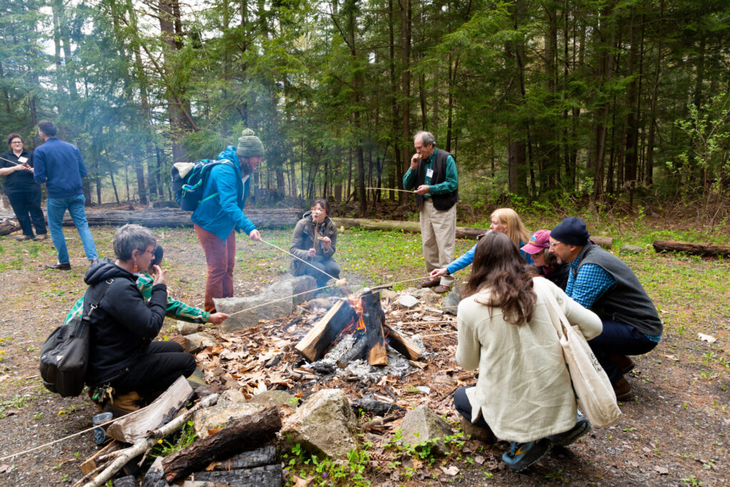 A group of people sit around a campfire talking and roasting marshmallows during the daytime